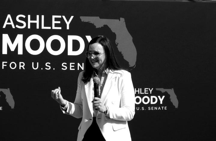 Ashley Moody holding a microphone in front of a Moody for U.S. Senate banner at Crabby Bill's in Indian Rocks Beach